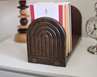 May include: A dark brown wooden bookend with an arched design, holding a stack of books. The books have colorful spines and a Penguin English Library cover. A decorative clock and lamp are in the background.