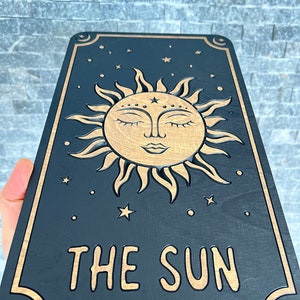 K&ouml;nnte beinhalten: Eine schwarze Tarotkarte aus Holz mit einem goldenen Sonnendesign, geschlossenen Augen, Sternen und Monden. Die Worte "THE SUN" sind in Gold graviert. Die Karte hat eine rechteckige Form. Das Design ist himmlisch.