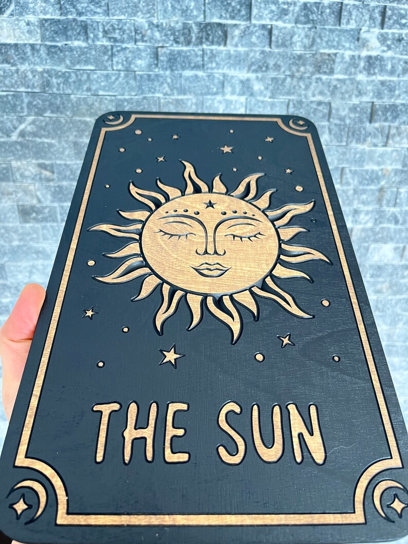 K&ouml;nnte beinhalten: Eine schwarze Tarotkarte aus Holz mit einem goldenen Sonnendesign, geschlossenen Augen, Sternen und Monden. Die Worte "THE SUN" sind in Gold graviert. Die Karte hat eine rechteckige Form. Das Design ist himmlisch.