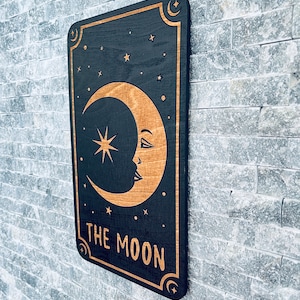 K&ouml;nnte beinhalten: Wandkunst aus Holz mit schwarzem Hintergrund, goldenem Halbmond, Sternen und dem Text "THE MOON". Das Design ist mit goldenen Akzenten und Halbmonddetails umrahmt. Die Kunst ist an einer grauen Backsteinmauer befestigt.