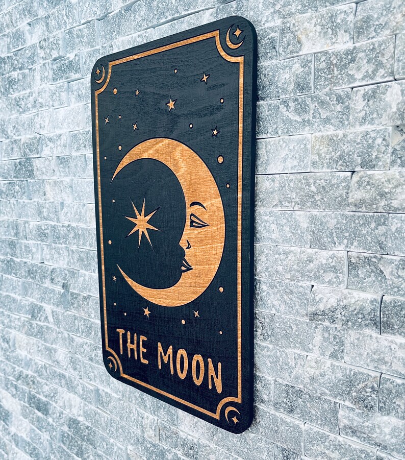 K&ouml;nnte beinhalten: Wandkunst aus Holz mit schwarzem Hintergrund, goldenem Halbmond, Sternen und dem Text "THE MOON". Das Design ist mit goldenen Akzenten und Halbmonddetails umrahmt. Die Kunst ist an einer grauen Backsteinmauer befestigt.
