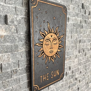K&ouml;nnte beinhalten: Wanddekoration aus Holz mit einem Tarotkarten-Design. Die schwarze Karte hat einen goldenen Rand und eine detaillierte Illustration der Sonne mit geschlossenen Augen. Der Text "THE SUN" steht unten, mit Sternen und Monden im Hintergrund.