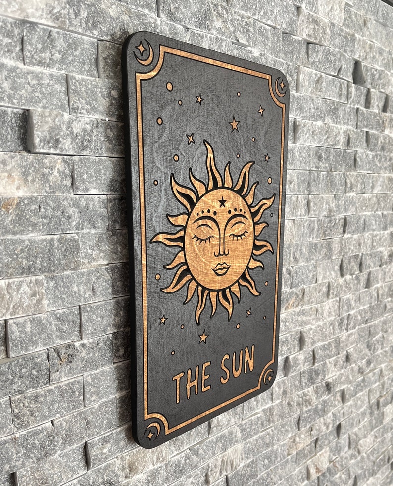 K&ouml;nnte beinhalten: Wanddekoration aus Holz mit einem Tarotkarten-Design. Die schwarze Karte hat einen goldenen Rand und eine detaillierte Illustration der Sonne mit geschlossenen Augen. Der Text "THE SUN" steht unten, mit Sternen und Monden im Hintergrund.