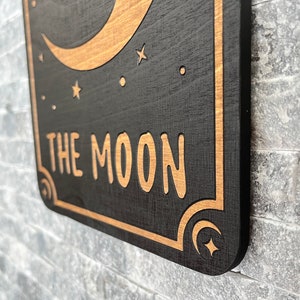 K&ouml;nnte beinhalten: Ein Holzschild mit schwarzem Hintergrund und goldfarbenen Akzenten. Das Schild zeigt einen Halbmond, Sterne und die Worte "THE MOON" in einer stilisierten Schriftart. Die R&auml;nder des Schildes sind ebenfalls goldfarben.