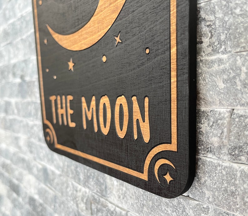 K&ouml;nnte beinhalten: Ein Holzschild mit schwarzem Hintergrund und goldfarbenen Akzenten. Das Schild zeigt einen Halbmond, Sterne und die Worte "THE MOON" in einer stilisierten Schriftart. Die R&auml;nder des Schildes sind ebenfalls goldfarben.