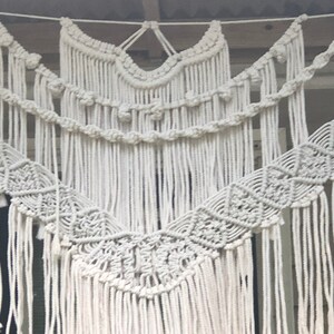 May include: A white macrame wall hanging with a geometric pattern and fringe. The hanging is made of cotton cord and has a bohemian style.