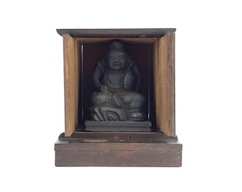 May include: A dark wooden shrine with a small, black, carved statue of a seated figure inside. The shrine has a hinged door on each side.