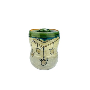 May include: A small ceramic jar with a green and blue lid. The jar has a white background with a black geometric design and hanging ornaments.