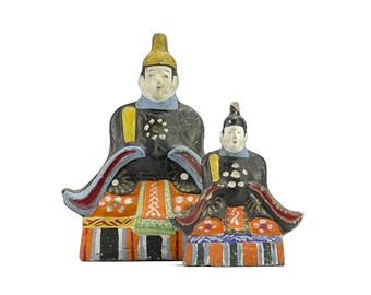 May include: Two traditional Japanese dolls, one larger than the other, are dressed in elaborate costumes with intricate details. The larger doll is wearing a black and gold robe with a red and white patterned skirt. The smaller doll is wearing a black and red robe with a red and white patterned skirt. Both dolls have painted faces and black hair.