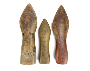 May include: Three antique wooden tools with pointed tops and rounded bases. The tools vary in size and color, with shades of brown and tan. The central tool is smaller and lighter in color than the two flanking it.
