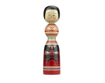 May include: A traditional Japanese wooden doll with a red, white, and blue striped body. The doll has a painted face with a black hair and a red and black demon face on the lower part of the body.