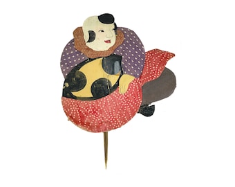 May include: A colorful paper cutout of a person holding a yellow and black object. The person is wearing a purple and white patterned shirt and a red and white patterned skirt. The cutout is attached to a wooden stick.
