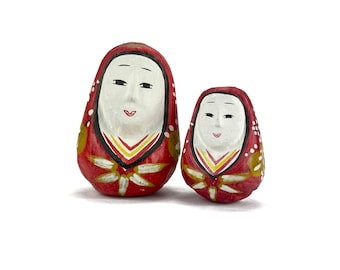 May include: Two Japanese Daruma dolls, one large and one small, stand side-by-side. The dolls are painted red with white faces, black eyes, and small red mouths. The larger doll is approximately 4 inches tall, while the smaller one is about 3 inches tall.