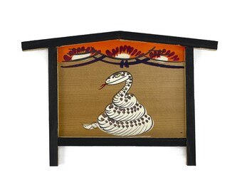 May include: Wooden sign featuring a stylized snake illustration. The snake is white with black spots and a red tongue, set against a light brown background. The sign has a black frame and a decorative orange and red top.
