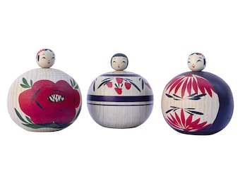 May include: Three wooden Japanese dolls with painted faces and colorful floral designs. The dolls are round and have a traditional Japanese style.