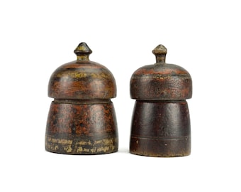 May include: Two antique wooden salt and pepper shakers. The shakers have a dark brown base with a lighter brown and gold top. The tops are rounded with a small, pointed finial. The wood has a distressed, aged appearance.