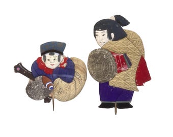 May include: Two vintage Japanese paper dolls. One doll is seated and wearing a blue hat, blue jacket, and red collar. The other doll is standing and wearing a tan kimono with red accents and a purple skirt. Both dolls have detailed facial features.