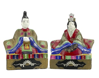 May include: A pair of traditional Japanese dolls, a male and a female, dressed in elaborate colorful robes and seated on platforms with intricate designs. The dolls are made of painted paper mache and are a popular decorative item for the Hinamatsuri festival.