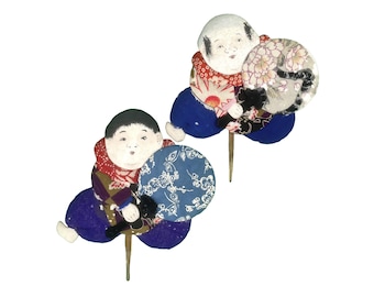 May include: Two small, hand-painted, fabric dolls with round faces and black hair. Each doll is holding a round fan with a floral design. The dolls are wearing red, blue, and white patterned clothing.