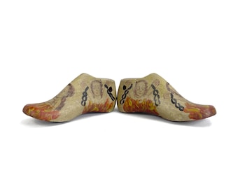 May include: A pair of antique wooden shoe forms. Each form is painted with a design featuring a face, flames, and chains. The forms are beige with red, orange, and black accents. The shoe forms are displayed on a white background.