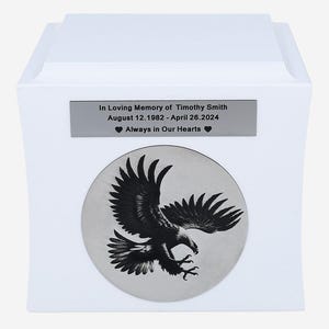 K&ouml;nnte beinhalten: Wei&szlig;e Urne mit einer silbernen Plakette mit der Aufschrift "In Loving Memory of Timothy Smith 12. August 1982 - 26. April 2024 Always in Our Hearts". Die Urne hat au&szlig;erdem einen silbernen Kreis mit einem Schwarzwei&szlig;bild eines schwebenden Adlers.