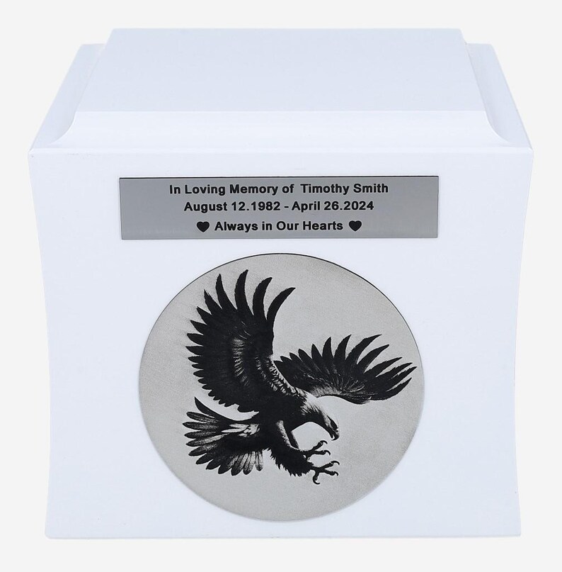 K&ouml;nnte beinhalten: Wei&szlig;e Urne mit einer silbernen Plakette mit der Aufschrift "In Loving Memory of Timothy Smith 12. August 1982 - 26. April 2024 Always in Our Hearts". Die Urne hat au&szlig;erdem einen silbernen Kreis mit einem Schwarzwei&szlig;bild eines schwebenden Adlers.