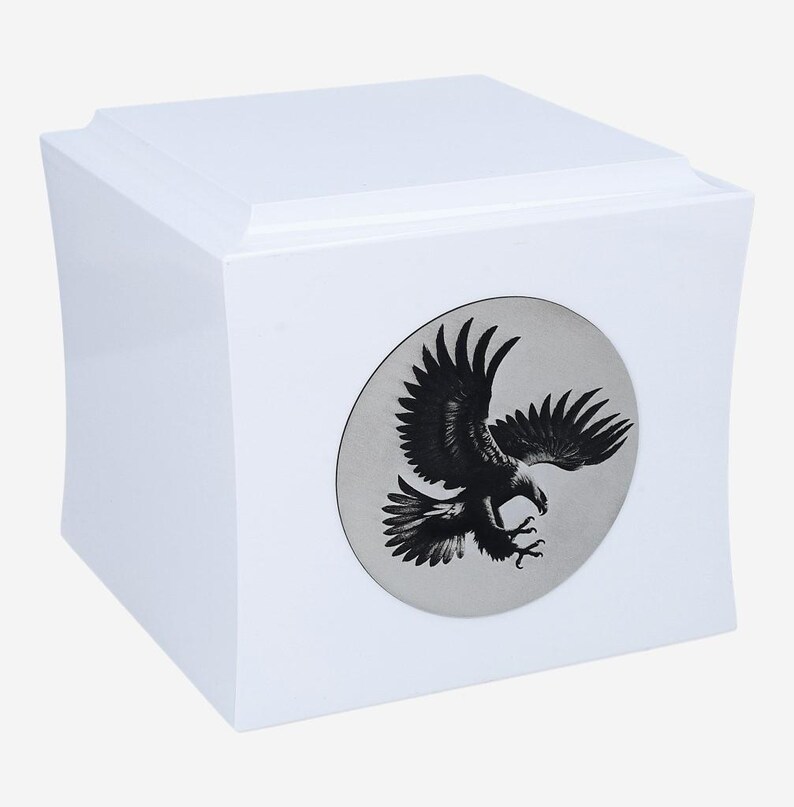 K&ouml;nnte beinhalten: Wei&szlig;e Urne f&uuml;r die Aschebestattung mit einem silbernen runden Emblem, das eine schwarze Silhouette eines Adlers im Flug zeigt.