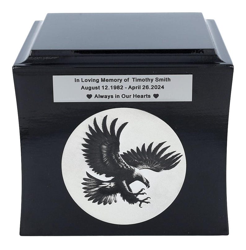K&ouml;nnte beinhalten: Schwarze Urne aus Holz mit einem wei&szlig;en Kreis, der ein schwarz-wei&szlig;es Adlermotiv zeigt. Die Urne hat eine silberne Plakette mit dem Text "In Loving Memory of Timothy Smith August 12.1982 - April 26.2024 Always in Our Hearts"