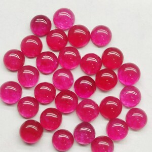 May include: A collection of round, polished gemstones in varying shades of red and pink. The gemstones are arranged in a scattered pattern, each with a smooth, reflective surface. The overall effect is a vibrant display of color and light.