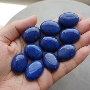 Pode incluir: Uma coleção de dez pedras preciosas ovais de lápis-lazúli polido. As pedras são de um azul profundo e vibrante com manchas douradas, seguradas em uma mão. As pedras têm uma superfície lisa e brilhante. Medem cerca de 2-3 cm.