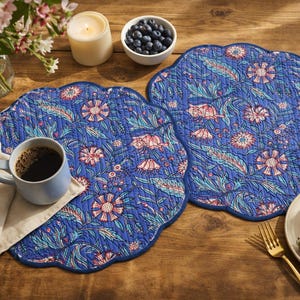 Puede incluir: Dos manteles individuales azules con estampado floral y bordes festoneados sobre una mesa de madera. Una taza de café azul claro, un cuenco de arándanos, una vela y una porción de pastel con fresas también están sobre la mesa. Hay una cuchara y un tenedor dorados.