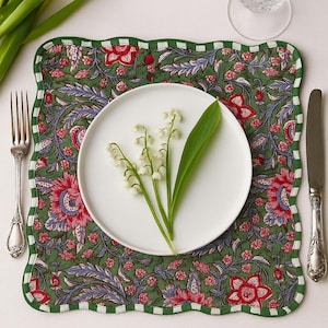 Peut inclure: Une assiette blanche ornée de fleurs de muguet repose sur un set de table vert à motifs floraux et bordure festonnée. Une fourchette, un couteau et un verre d'eau complètent la mise en place.