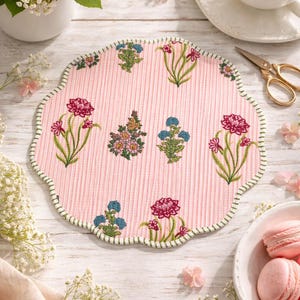 May include: A pink, scalloped-edge placemat with a floral pattern. The placemat features embroidered flowers in shades of pink, blue, and yellow, with a green border. The background is a light pink with vertical stripes. The image also includes a cup of tea, flowers, and macarons.