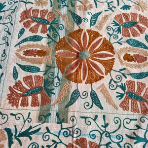 May include: Cream-colored textile with an embroidered floral pattern. The design features large, stylized flowers in shades of orange and brown, with teal and green accents. Intricate vines and swirls complete the design.