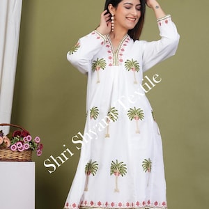 May include: White cotton dress with a V-neckline and long sleeves. The dress features a green and pink palm tree print and a pink and gold trim around the neckline and hem. The dress is knee-length and has a loose, flowing fit. "Shri Shyam Textile" is printed on the dress.
