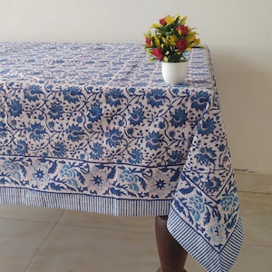 Op de afbeelding: Een wit tafelkleed met een blauw bloemenpatroon en een blauw-wit gestreepte rand. Een kleine witte vaas met gele en rode bloemen staat op het tafelkleed.