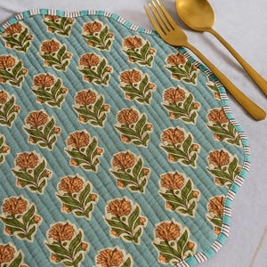 Può includere: Un sottopentola rotondo e trapuntato con sfondo azzurro e motivo floreale ripetuto arancione e verde. Il sottopentola ha un bordo smerlato con un bordo a righe. Una forchetta e un cucchiaio dorati sono posizionati sul sottopentola.