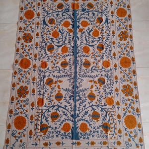 May include: A beige embroidered tapestry featuring a stylized tree of life design with blue and orange floral motifs. The tree is surrounded by a border of smaller floral designs.