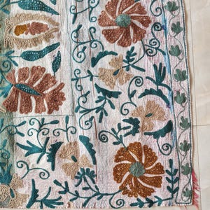 May include: Close-up of a white textile with intricate embroidery. The design features large brown and tan flowers, teal leaves, and swirling vines. The edges are trimmed with a teal and green border, and a fringe of pink threads.