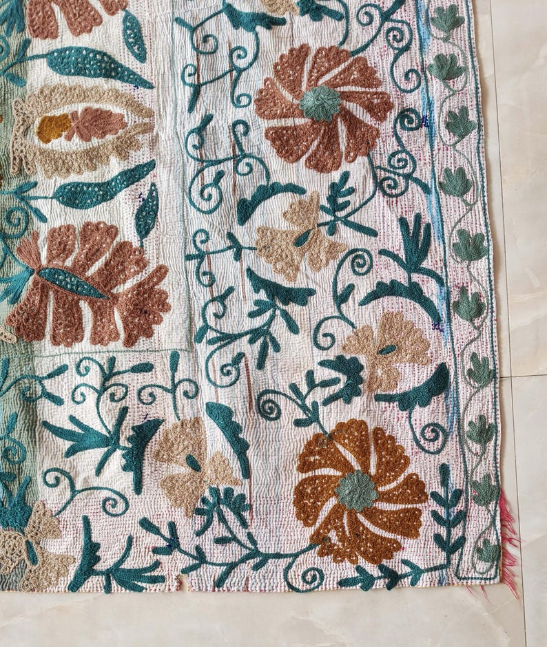 May include: Close-up of a white textile with intricate embroidery. The design features large brown and tan flowers, teal leaves, and swirling vines. The edges are trimmed with a teal and green border, and a fringe of pink threads.