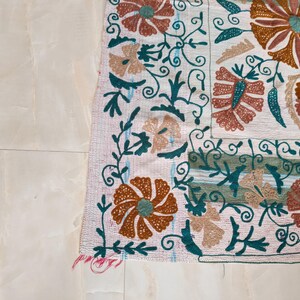 May include: Close-up of a section of a white textile with intricate floral embroidery. The design features large, stylized flowers in shades of orange, teal, and beige, with swirling green vines and leaves. The edges are finished with a fringed border.