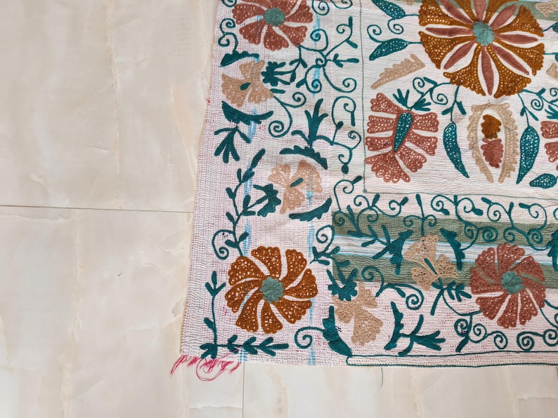 May include: Close-up of a section of a white textile with intricate floral embroidery. The design features large, stylized flowers in shades of orange, teal, and beige, with swirling green vines and leaves. The edges are finished with a fringed border.