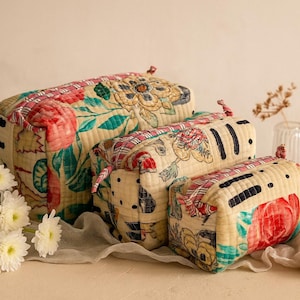 May include: Three quilted cosmetic bags in different sizes, patterned with flowers in red, pink, and teal on a cream base. Each bag has a zip closure and a small loop. A lipstick and a small gold box are placed beside the bags.