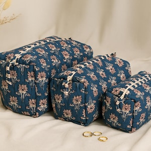 Op de afbeelding: Drie blauwe cosmetische tassen met bloemmotief in verschillende maten. De tassen hebben een witte rits en zijn gerangschikt op een wit oppervlak. Een witte vaas met gedroogde bloemen staat op de achtergrond. Gouden ringen staan op de voorgrond.
