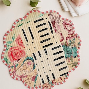 May include: A floral patterned, quilted placemat with a scalloped edge. The placemat features a patchwork design with cream, red, and black accents. The edge is trimmed with a red and white striped fabric. A rose and greenery are in the background.