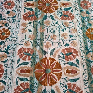 May include: Cream-colored textile with intricate floral embroidery. The design features large, stylized flowers in shades of orange, brown, and teal, surrounded by delicate vines and leaves. The embroidery is detailed and creates a textured surface.
