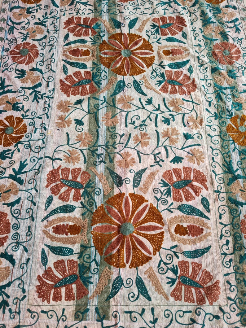 May include: Cream-colored textile with intricate floral embroidery. The design features large, stylized flowers in shades of orange, brown, and teal, surrounded by delicate vines and leaves. The embroidery is detailed and creates a textured surface.