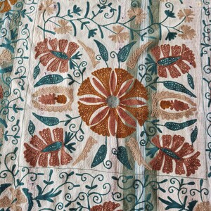 May include: Close-up of a cream-colored textile with intricate embroidery. The design features large, stylized flowers in shades of brown and peach, with teal and beige accents. The floral pattern is set against a background of scrolling vines.
