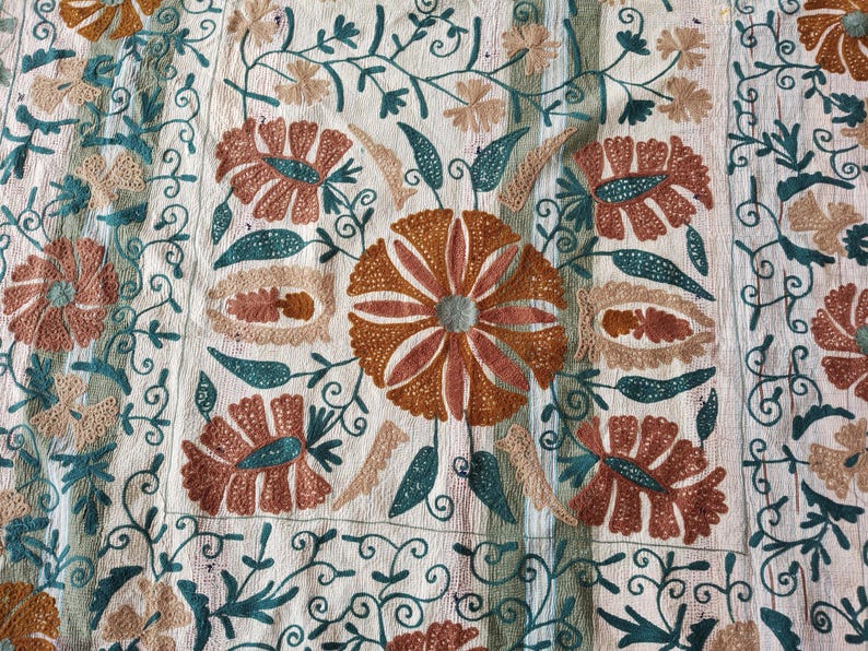May include: Close-up of a cream-colored textile with intricate embroidery. The design features large, stylized flowers in shades of brown and peach, with teal and beige accents. The floral pattern is set against a background of scrolling vines.
