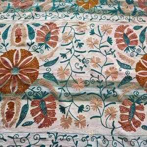 May include: Close-up of a cream-colored textile with intricate embroidery. The design features large, stylized flowers in shades of orange and brown, with teal accents. Delicate vines and smaller flowers fill the spaces, creating a detailed pattern.